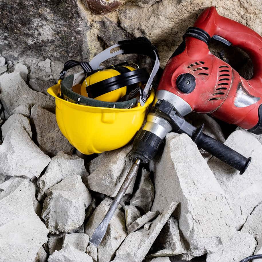 Bauarbeiterhelm und Schlagbohrer auf Schutthaufen. Demontage von nicht-tragenden Bauteilen: Zunächst werden alle nicht tragenden Wände, Decken, Bodenbeläge und Inneninstallationen wie Türen, Fenster, Heizungen, sanitäre Anlagen, Elektroleitungen und Lüftungsanlagen entfernt.