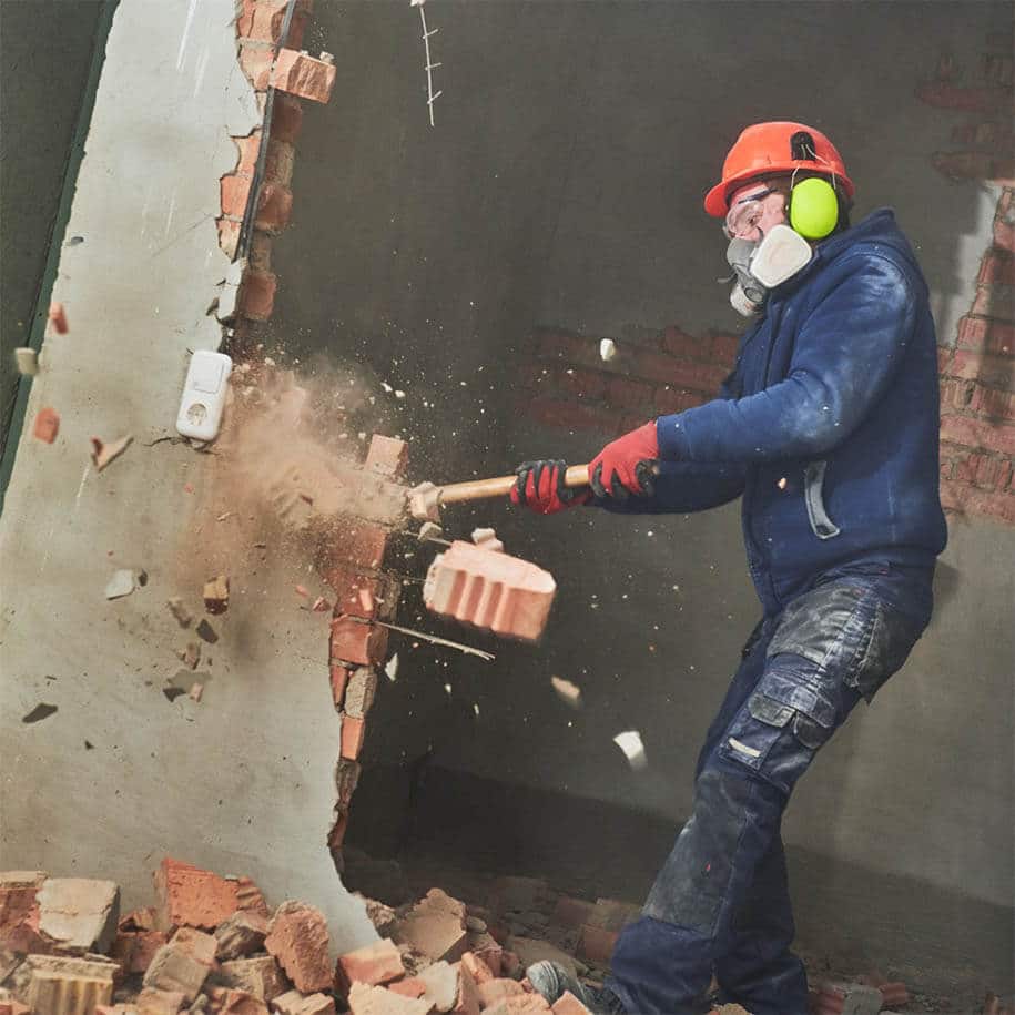 Arbeiter beim Abriss einer Wand mit dem Vorschlaghammer. Entfernung von Einbauten: Fest installierte Einbauten wie Küchen oder Schränke werden abgebaut. Entsorgung: Gefährliche Abfälle müssen sicher entsorgt werden, während andere Materialien möglicherweise recycelt werden können.